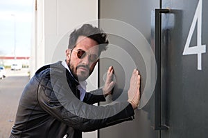 handsome young man with beard, sculpted body and sunglasses is leaning with both hands on the grey door of his garage looking at