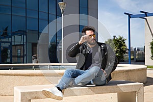 Handsome young man with beard, sunglasses, leather jacket and jeans, sitting on a stone bench, relaxed. Concept beauty, fashion,