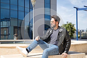 Handsome young man with beard, sunglasses, leather jacket and jeans, sitting on a stone bench, relaxed. Concept beauty, fashion,