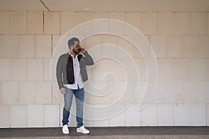 Handsome young man with beard, sunglasses, leather jacket, white shirt and jeans, next to a white wall, talking on his cell phone