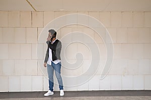 Handsome young man with beard, sunglasses, leather jacket, white shirt and jeans, next to a white wall, talking on his cell phone