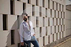 Handsome young man with beard, sunglasses and open white shirt, leaning against a wall of white stone blocks, smiling in sexy