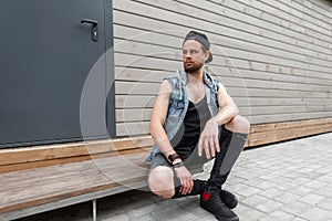 Handsome young man with a beard in a trendy denim vest in a T-shirt in stylish ripped jeans in sneakers in a vintage cap sits