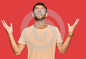 Handsome young man with beard wearing casual tshirt crazy and mad shouting and yelling with aggressive expression and arms raised