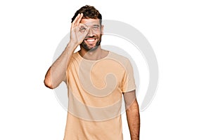 Handsome young man with beard wearing casual tshirt doing ok gesture with hand smiling, eye looking through fingers with happy