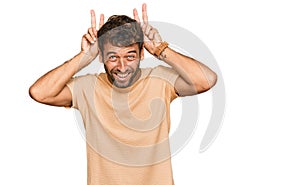 Handsome young man with beard wearing casual tshirt posing funny and crazy with fingers on head as bunny ears, smiling cheerful