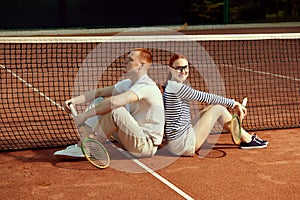 Handsome young man and beautiful woman in casual, stylish clothes posing with tennis racket on court on daytime
