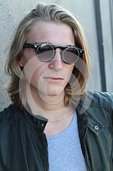 Handsome young man with blond long hair and sunglasses isolated on grey background. Fashion studio shot