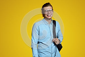 Handsome young man in blue shirt and eyeglasses holding backpack and smiling at camera on yellow backdrop