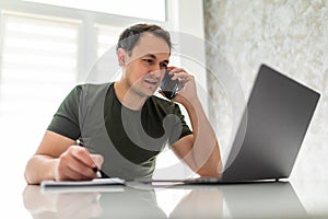 Handsome young man calculate bills make notice using laptop and talking on cellphone in kitchen