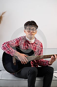 Handsome young man in casual clothes is looking at camera while playing guitar at home.