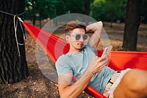 Handsome young man in casual clothes resting in the woods lying in a hammock on a tree and using a smartphone with a serious face