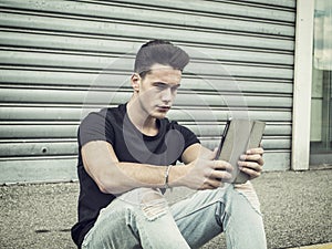 Young man using tablet PC sitting on street curb