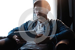 Handsome young man in casual clothes using a digital tablet and headphones and smiling while sitting on the chair.