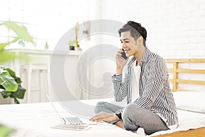 Handsome young man in casual clothes is working at home while sitting on the bed,with a laptop in bedroom,and talking on the phone