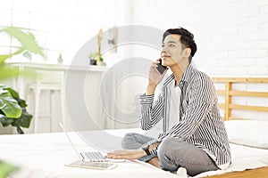 Handsome young man in casual clothes and working at home while sitting on the bed,with a laptop and talking on phone in bedroom