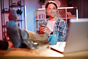 Handsome young man in checkered shirt put his feet on table with modern smartphone in hands.