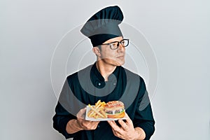 Handsome young man chef holding burger with fries smiling looking to the side and staring away thinking