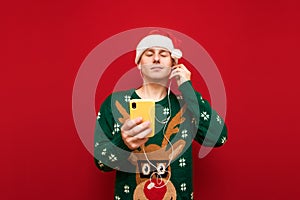 Handsome young man in a Christmas hat and sweater listening to music in headphones with his eyes closed and holding a smartphone