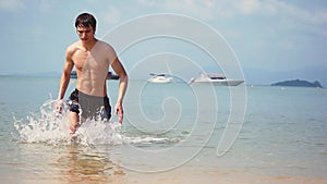 Handsome young man comes out of the sea beach trunks swimmer wet athletic body muscular brawny satisfied happy summer