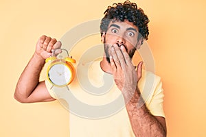 Handsome young man with curly hair and bear holding alarm clock covering mouth with hand, shocked and afraid for mistake