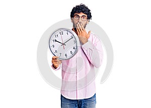 Handsome young man with curly hair and bear holding big clock covering mouth with hand, shocked and afraid for mistake