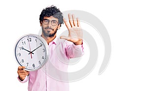 Handsome young man with curly hair and bear holding big clock with open hand doing stop sign with serious and confident