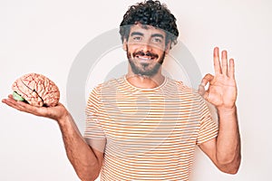 Handsome young man with curly hair and bear holding brain as mental health concept doing ok sign with fingers, smiling friendly