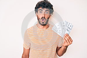 Handsome young man with curly hair and bear holding money bag with dollar symbol scared and amazed with open mouth for surprise,