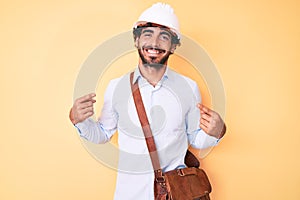 Handsome young man with curly hair and bear wearing architect hardhat looking confident with smile on face, pointing oneself with