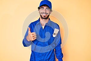 Handsome young man with curly hair and bear wearing builder jumpsuit uniform doing happy thumbs up gesture with hand