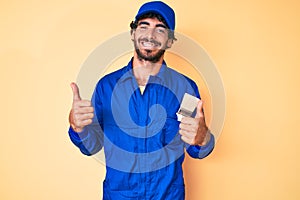 Handsome young man with curly hair and bear wearing builder jumpsuit uniform success sign doing positive gesture with hand, thumbs