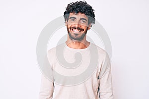 Handsome young man with curly hair and bear wearing casual winter sweater with a happy and cool smile on face