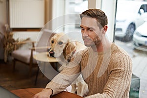 Handsome young man, digital nomad sitting with his dog in a pet-friendly cafe, working on laptop while a golden