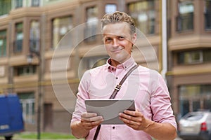 Handsome young man with digital tablet in hands stand outdoors in front urban office building wearing casual pink shirt