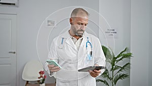 Handsome young man doctor, deeply concentrated, working online, typing a message on touchpad and cellphone in clinic\'s waiting