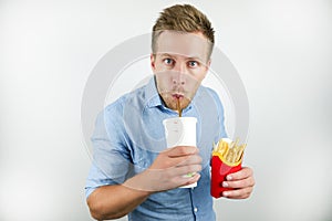 Handsome young man drinks soda from papper cup and holds fries from fast food restaurant on isolated white background