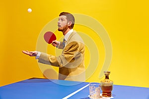 Handsome young man in elegant suit player table tennis against bright yellow background. Whiskey glass on table