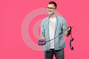 handsome young man in eyeglasses holding purple rotary phone and smiling at camera isolated