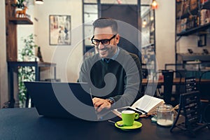 Handsome young man in eyeglasses is using laptop and smiling while sitting in cafe