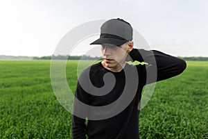 Handsome young man in a fashionable black baseball cap in a vintage black shirt posing among green grass in a field