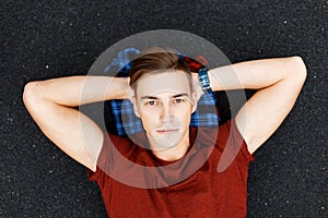 Handsome young man in a fashionable red t-shirt with a stylish hairstyle lies on a black asphalt on a plaid shirt. Cute guy