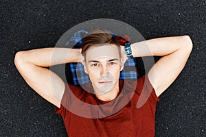 Handsome young man in a fashionable red t-shirt with a stylish hairstyle lies on a black asphalt on a plaid shirt. Cute guy is