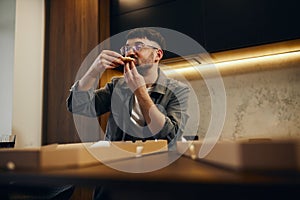 A handsome young man in glasses is enjoying pizza in his kitchen. Unbelievably enjoys the taste