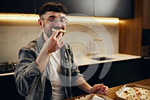 A handsome young man in glasses is enjoying pizza in his kitchen. Unbelievably enjoys the taste