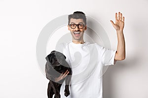 Handsome young man in glasses holding his black pug and waving hand, guy saying hello while carry dog with one arm