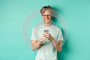 Handsome young man in glasses with red messy hair reading message on mobile phone, smiling and looking at screen