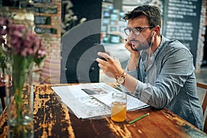 Handsome young man with glasses sitting in cafe using smarphone and listening with earphones