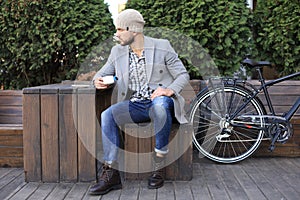 Handsome young man in grey coat and hat sitting on a bench relaxed drinking coffee and thinking near his bicycle