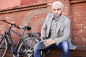 Handsome young man in grey coat and hat sitting on a bench relaxed drinking coffee and thinking near his bicycle